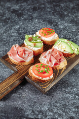 Assorted Italian bruschette snack on a wooden chopping board on a dark textured table. Mini sandwich mix. Angle view