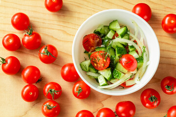 Cucumber, cherry tomatoes and onion salad in a deep white bowl on a light wooden Board. Next to the bowl is a lot of red cherry tomatoes