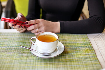 Close-up of a mobile phone in the hands of a woman, texting and sitting over a cup of tea. Bright morning in a cafe