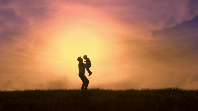 Against the background of the bright orange sunset sky, the silhouette of the father hugs the silhouette of his daughter, circles her and throws her up into the sky. Overall plan.