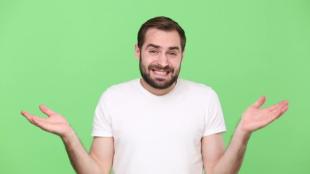 Bearded fun confused young guy 20s in white t-shirt isolated on green chroma key background studio. People sincere emotions lifestyle concept. Looking at camera spreading hands say oops i am sorry