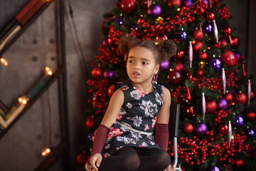 Beautiful kid girl 5-6 year old wearing stylish dress sitting in armchair over Christmas tree in room. Looking at camera. Holiday season.