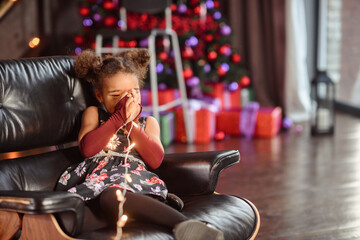 Beautiful kid girl 5-6 year old wearing stylish dress sitting in armchair over Christmas tree in room. Looking at camera. Holiday season.