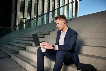 Fototapeta premium Young businessman sits on the stairs between high-rise buildings and is working with his laptop.