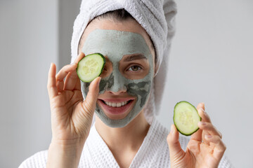 Young smiling woman with cosmetic clay facial mask on face hold fresh cucumber slices.