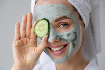 Young woman with grey  cosmetic natural clay mask on face skin and fresh green cucumber slice on...