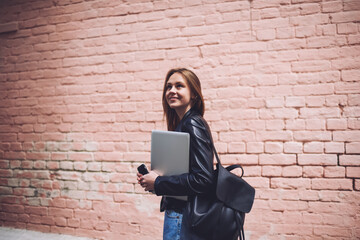 Dreamy hipster girl walking with stylish backpack and laptop device in hand and looking up.