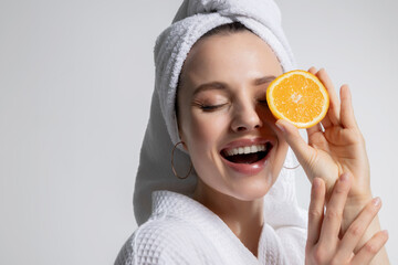 Smiling happy young beautiful girl in bathrobe and head towel holding orange citrus fruit round...