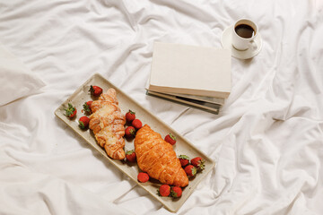 Croissants with strawberry, coffee cup, and open book on a white bedsheet. Background with space. Good morning home concept. Delicious breakfast in bed. Top view. Flat lay