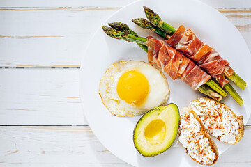 fried asparagus wrapped in jamon, fried eggs, bruschetta with soft cheese and avocado on a white plate