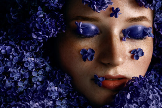 Close up beauty portrait of beautiful redhead woman with freckled skin, wet blue eyes makeup posing in lilac flowers. Close up portrait