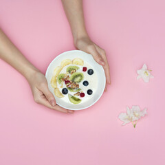 Female hands holding a bowl of smoothies with superfoods. Yogurt. blueberries, kiwi, almonds, banana, nuts. Top view on a pink background.