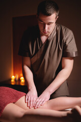 Close-up professional calf muscle massage to a female client by a male physiotherapist in a massage parlor in a dark office against the background of burning candles