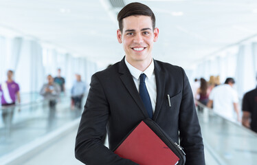 Portrait of confident smiling young businessman in a suit and tie looking into the camera while standing in front of crowded corporate terminal moving walkways.