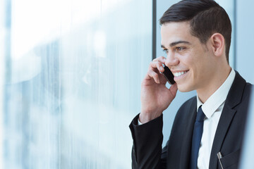 Smiling businessman wearing suit and tie speaking on mobile phone with client at work near the office window.

