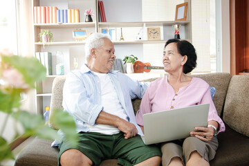 An elderly Asian couple sitting on a sofa watching media online on a laptop In the living room at home Both are happy in life after retirement. Concept of health insurance, Social distance