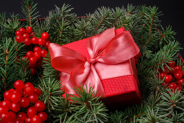 Fototapeta na wymiar New Year Christmas Xmas holiday celebration red present gift box with satin pink bow, immersed in the needles of a Christmas tree decorated with red berries. Dark festive composition.
