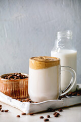 Dalgona frothy coffee trend korean drink milk latte with coffee foam in glass mug on ceramic tray with ingredients on white marble table.