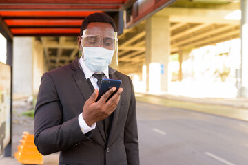 African businessman with mask and face shield using phone at the bus stop in the city outdoors