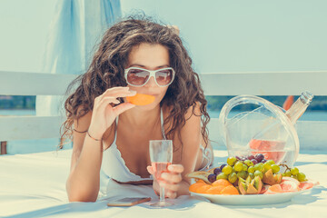 woman  holding champagne glass, eating orange on a sun bed