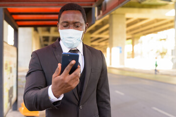 African businessman with mask using phone while waiting at the bus stop