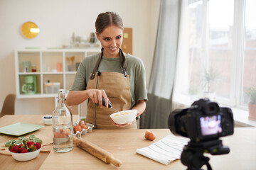 Smiling young blogger beating the eggs in the bowl in her hands and showing it to the camera while...
