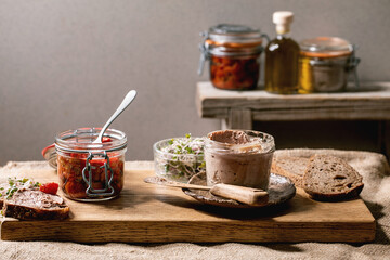 Glass jar of homemade chicken liver pate with sliced rye bread, sun-dried tomatoes and green sprout...