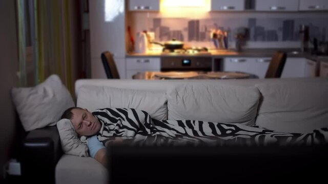 Tired young man lying on a sofa covered with a plaid, watching TV, camera movement