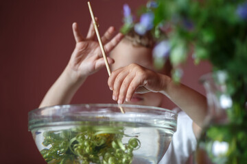 with his left hand the boy holds a wooden stick, the fingers of his right hand are preparing to take it. in the foreground is a transparent glass bowl with water, dandelions float in it