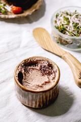 Ceramic bowl of homemade chicken liver pate with wooden knife, sliced rye bread, sun-dried tomatoes and green sprout salad on white linen table cloth. Home breakfast or appetizer