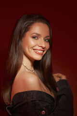 Happy smiling woman with long straight brunette hair. Girl wearing necklace and earrings, bra and jacket posing against red bakcground. Fashion style portrait