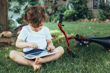 Child sitting on the grass with a tablet and red bike.