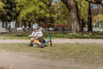Fototapeta premium A cute little boy in camouflage beanie is sitting on his runbike in the park. He is holding a stick and examining it.