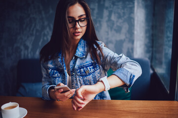 Emotional young woman in eyeglasses looking at alarm watch sitting in coffee shop interior.