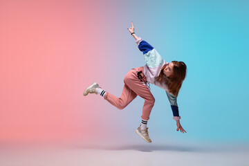 In jump. Beautiful girl dancing hip-hop in stylish clothes on colorful gradient background at dance...