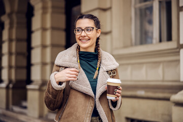 Young smiling college girl standing in front of her university, holding disposable cup with coffee...
