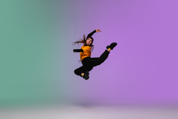 Naklejka na ściany i meble In jump. Beautiful girl dancing hip-hop in stylish clothes on colorful gradient background at dance hall in neon light. Youth culture, movement, style and fashion, action. Fashionable bright portrait.
