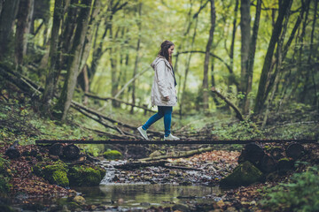 Side view of a young woman in a grey jacket and jeans walking over a wooden  bridge over water in an enchanted mystical forest