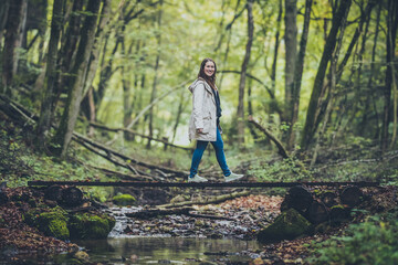 Side view of a young woman in a grey jacket and jeans walking over a wooden  bridge over water in an enchanted mystical forest