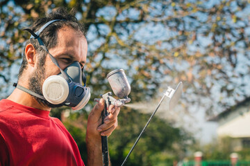 Amateur painter in red sweater and face mask using a spray gun to paint a plastic object or part in a home garden. Profile view of a painter with a small spray gun.