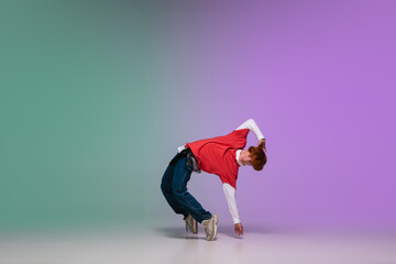 Beautiful redhead boy dancing hip-hop in stylish clothes on colorful gradient background at dance hall in neon light. Youth culture, movement, style and fashion, action. Fashionable bright portrait.