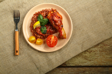 Chicken legs with red rice in tomato-garlic sauce on a plate on a wooden table on a linen tablecloth and fork.