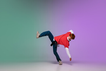 Fototapeta premium Beautiful redhead boy dancing hip-hop in stylish clothes on colorful gradient background at dance hall in neon light. Youth culture, movement, style and fashion, action. Fashionable bright portrait.