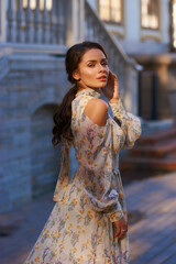 Fototapeta premium Pretty smiling young woman with long brunette hair wearing fashionable floral dress standing on street against balustrade on background. Happy female model in elegant stylish outfit posing outdoors.