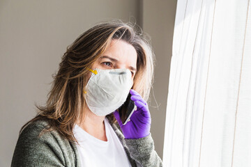 girl at home with sanitary mask and latex gloves, using a mobile phone, next to the bedroom window on a sunny day. Concept: Stay home. Pandemic, coronavirus, covid 19. isolation communication,
