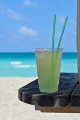 Cooled cocktail on a wooden table on Varadero beach in Cuba, the drink in a plastic mug with straws, blurred turquoise caribbean sea in the background, a sunny day