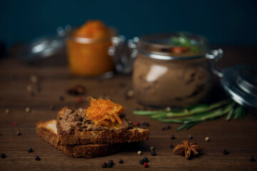 A slice of bread with poultry pate and orange gam, glass jar with poultry pate, decorated with almonds and rosemary, and jar with orange jam on a wooden table