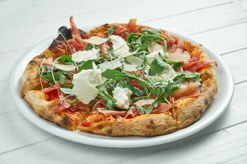 Appetizing homemade pizza with jamon, parmesan, arugula and straccella cheese on a white plate on a white wooden background