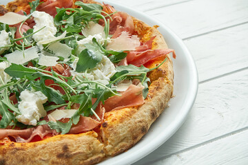 Appetizing homemade pizza with jamon, parmesan, arugula and straccella cheese on a white plate on a white wooden background