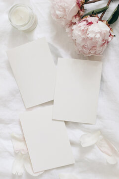Wedding stationery, still life composition. Greeting cards mockup scene. Blank sheets of paper and pink peony flowers on white linen table cloth. Vintage feminine styled photo, flat lay, top view,